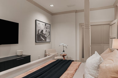 Cozy modern bedroom interior in neutral tones with a layered beige-and-black bed, plush pillows, wall-mounted TV, dark console, beige armchair and side table with a white orchid, framed black-and-white photo, and soft recessed lighting.