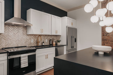 Modern kitchen with white shaker cabinets, herringbone tile backsplash, stainless steel appliances, matte black island with decorative bowl, gold faucet and globe pendant lights