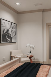 Cozy modern bedroom corner with a beige armchair, small side table holding a white orchid, a framed black-and-white piano photograph on the wall, neutral walls and soft bedding with a dark throw.
