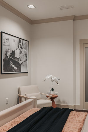 Cozy modern bedroom corner with a beige armchair, small side table holding a white orchid, a framed black-and-white piano photograph on the wall, neutral walls and soft bedding with a dark throw.