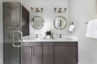 Contemporary master bathroom with double sink vanity — dark gray cabinets, white quartz countertop, two round gold-framed mirrors, dual modern faucets, potted plant, glass shower door and towel rack