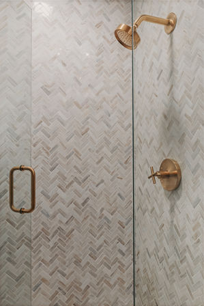 Modern shower with white marble herringbone tile, glass door, and brushed brass showerhead, handle, and control.