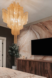 Gold fringe chandelier suspended above a modern bedroom with an abstract marble-style wall mural, wall-mounted TV, walnut media cabinet, potted eucalyptus and neutral bedding.