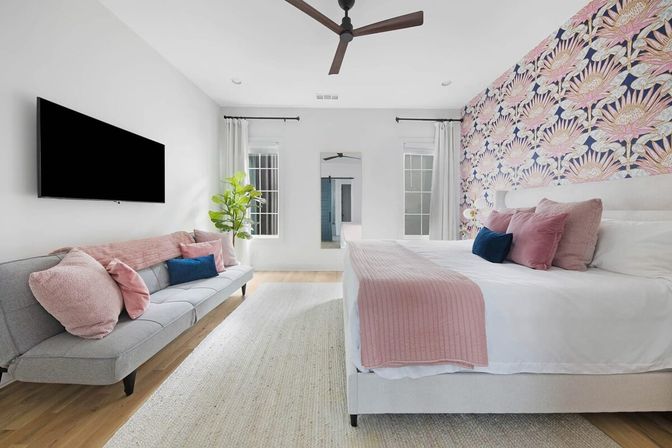 Bright modern bedroom interior with floral accent wallpaper, king bed with pink quilt and throw pillows, gray sofa with pink cushions, wall-mounted TV, ceiling fan, fiddle-leaf plant and hardwood floors.