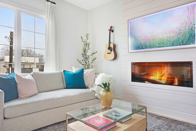 Bright contemporary living room with light-gray sofa and teal/blush pillows, glass coffee table with books and gold vase of white flowers, wall-mounted acoustic guitar, built-in linear fireplace with glowing flames beneath colorful artwork, large windows and potted plant.