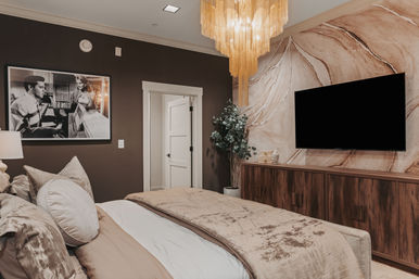 Cozy contemporary master bedroom interior with king bed in neutral linens, marble-effect accent wall, wall-mounted flat-screen TV above a wooden media console, gold tiered chandelier, potted plant, and framed vintage black-and-white photo.