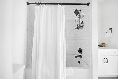 Bright minimalist white bathroom with subway-tiled bathtub, flowing white shower curtain, matte black showerhead and faucet, wall-mounted shower caddy and white vanity