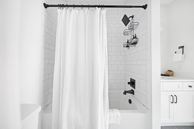 Bright minimalist white bathroom with subway-tiled bathtub, flowing white shower curtain, matte black showerhead and faucet, wall-mounted shower caddy and white vanity