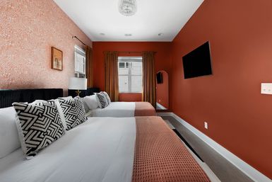 Modern bedroom with two beds, terracotta walls and floral accent wallpaper, black-and-white geometric throw pillows, white linens and textured rust throws, wall-mounted TV, window with gold curtains and arched floor mirror.