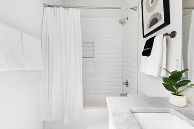Bright minimalist white bathroom with subway-tiled tub and shower curtain, marble vanity with sink, wall-mounted towels and a small potted plant.