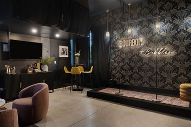Intimate industrial-chic cocktail lounge interior with a small performance stage, neon sign referencing bourbon and bubbles, velvet chairs, high bar table, and dark patterned wallpaper.