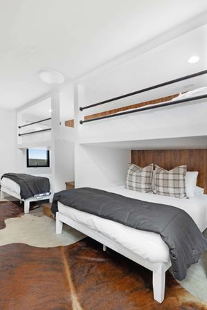 Bright modern bunk-room with built-in white loft beds, wood-paneled headboards, gray duvets and plaid pillows, black metal railings and brown cowhide rug