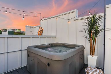 Rooftop hot tub on a private urban terrace at sunset, lit by string lights with a potted palm and rolled towels nearby.