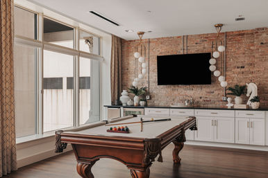 Stylish urban loft game room with a wooden pool table and racked billiard balls, large floor-to-ceiling windows, exposed brick wall, wall-mounted TV, globe pendant lights, white cabinetry and hardwood floors.