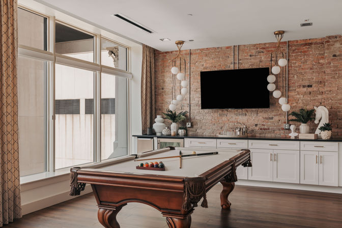 Stylish urban loft game room with a wooden pool table and racked billiard balls, large floor-to-ceiling windows, exposed brick wall, wall-mounted TV, globe pendant lights, white cabinetry and hardwood floors.
