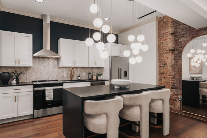 Sleek modern kitchen with white cabinets, stainless steel appliances, herringbone tile backsplash, black island with three plush bar stools, hanging globe pendant lights, exposed brick wall and hardwood floors.