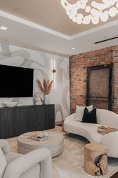 Modern urban loft living room with exposed brick wall, curved cream sofa, textured armchair, round stone coffee table, dark wood TV console and sculptural lighting.