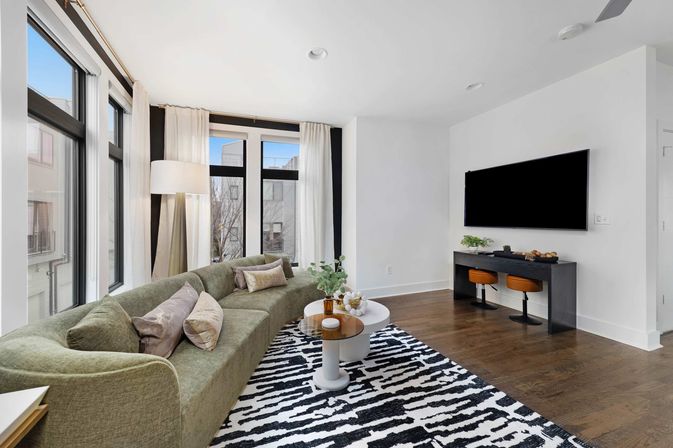 Bright modern city apartment living room with a curved olive-green sofa, patterned black-and-white rug, round nesting coffee tables, floor-to-ceiling windows with white curtains, wall-mounted flat-screen TV above a console with two orange stools, hardwood floors and a small potted plant.