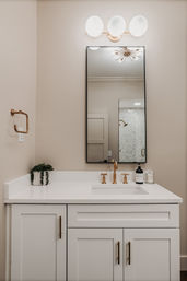 Sleek residential bathroom vanity with white quartz countertop, black-framed mirror, brushed-gold faucet and hardware, triple-globe wall sconce, small potted succulent, and neutral beige walls.