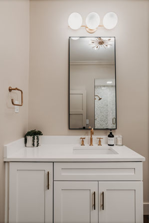 Sleek residential bathroom vanity with white quartz countertop, black-framed mirror, brushed-gold faucet and hardware, triple-globe wall sconce, small potted succulent, and neutral beige walls.