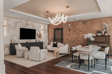 Open-plan urban loft living and dining room with exposed brick feature wall, curved cream sofa and boucle armchairs facing a wall-mounted TV, sculptural globe chandelier, dark wood sideboard and piano, modern dining table with upholstered chairs and white orchid centerpiece.
