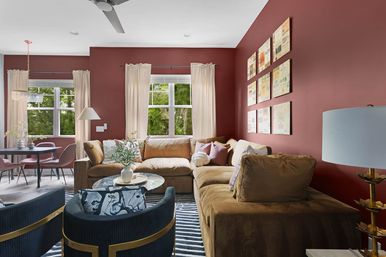 Cozy modern living room with deep red walls, large tan sectional, navy velvet armchairs with gold trim, striped rug and round marble coffee table with plant; cream curtains framing bright windows and an open-plan dining area with pink chairs and chandelier visible in the background.