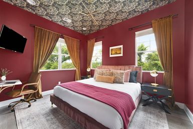 Contemporary bedroom with deep red walls and floral-patterned ceiling, pink velvet bed topped with textured pillows and magenta throw, gold curtains framing windows to leafy trees, brass lamps, black nightstands, wall TV and small desk with leather chair.