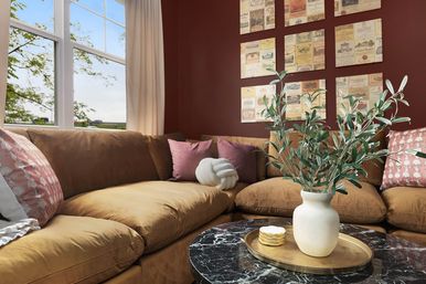 Cozy sunlit living room corner with caramel brown sectional sofa, burgundy accent wall gallery, marble coffee table with gold tray and ceramic vase of green branches, and pink patterned throw pillows by a window view.