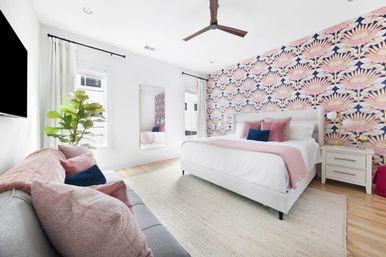 Bright modern bedroom with bold pink floral accent wall, crisp white bed with blush and navy pillows, fiddle‑leaf fig, full‑length mirror, light hardwood floors and ceiling fan.