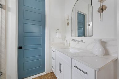 Bright modern coastal-style bathroom with a blue five-panel door, white shaker vanity, marble-look countertop, wall-mounted faucet, arched mirror, brass sconces and a decorative white vase.