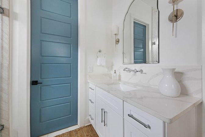 Bright modern coastal-style bathroom with a blue five-panel door, white shaker vanity, marble-look countertop, wall-mounted faucet, arched mirror, brass sconces and a decorative white vase.