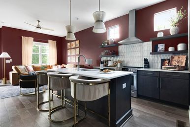 Open-concept modern kitchen and living room with deep burgundy walls, marble-topped island, gold-accent bar stools, stainless range hood, geometric tile backsplash, floating shelves and a cozy sofa by a sunlit window.