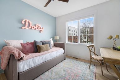Cozy pastel bedroom with gray daybed piled with pink and neutral pillows, a glowing pink marquee name sign on a pale blue accent wall, wooden desk and chair by a large window with city apartment views.