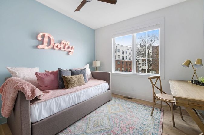 Cozy pastel bedroom with gray daybed piled with pink and neutral pillows, a glowing pink marquee name sign on a pale blue accent wall, wooden desk and chair by a large window with city apartment views.