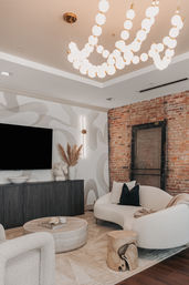 Cozy modern, loft-style living room with exposed brick accent wall, curved white sofa, black throw pillow, round stone coffee table, textured rug, wood stump side table, pampas grass vase, and sculptural globe-bulb chandelier
