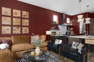 Modern open-concept apartment living room and kitchen with deep red accent wall, caramel velvet sofa, black velvet armchairs with patterned pillows, round black marble coffee table, zebra rug, and island with pendant lights and dark cabinetry