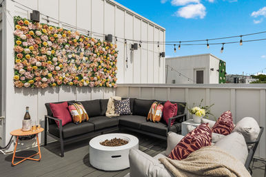 Sunny urban rooftop patio with a modern L-shaped black sectional, patterned pillows and throw, round white fire pit, large flower wall with neon sign, string lights overhead and a small side table with a bottle and glasses.