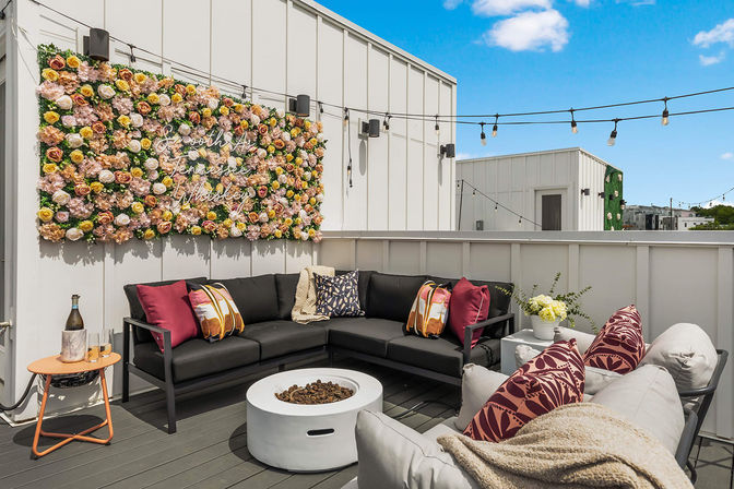 Sunny urban rooftop patio with a modern L-shaped black sectional, patterned pillows and throw, round white fire pit, large flower wall with neon sign, string lights overhead and a small side table with a bottle and glasses.