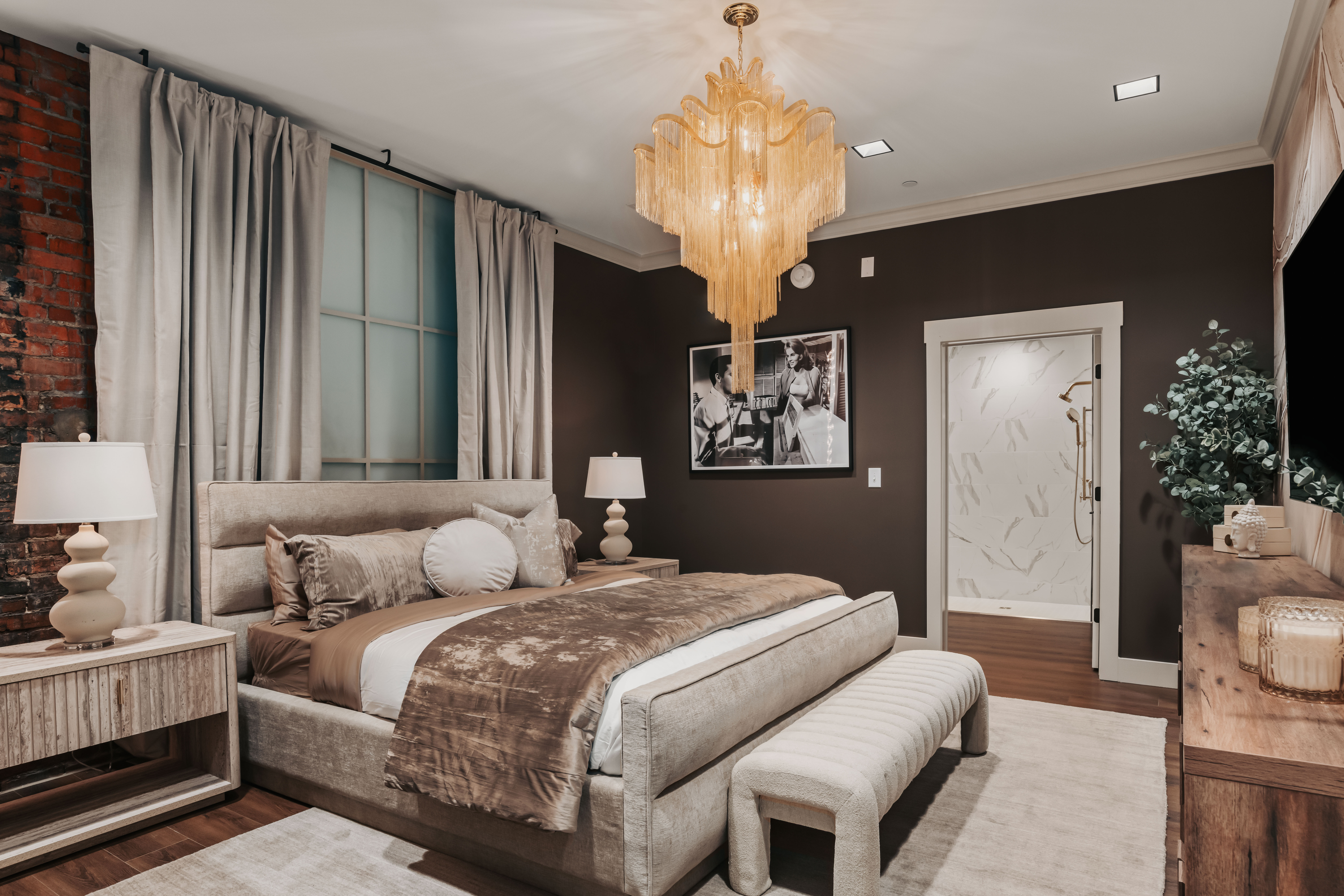 Luxurious modern bedroom interior with plush upholstered bed, gold fringe chandelier, exposed brick accent, neutral drapes, and en-suite marble shower.