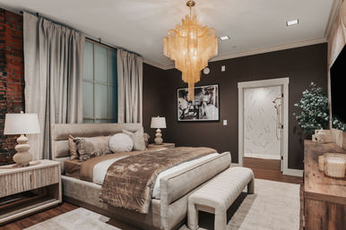 Luxurious modern bedroom interior with plush upholstered bed, gold fringe chandelier, exposed brick accent, neutral drapes, and en-suite marble shower.