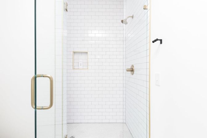 Bright modern bathroom shower with white subway tile walls, frameless glass door, brass handle and fixtures, and small white hex tile floor.