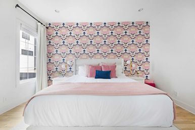 Sunlit modern bedroom in an urban apartment with white bedding, pink throw and pillows, navy accent pillow, pink-and-navy floral feature wall, white curtains and light hardwood floor.