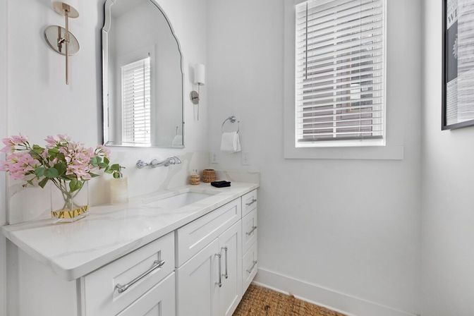 Sunlit modern white bathroom with marble countertop vanity, arched mirror, brass wall sconces, vase of pink flowers, window blinds and woven floor rug