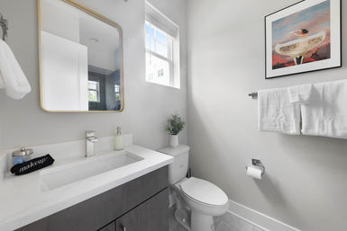 Bright modern small bathroom with white marble countertop and undermount sink, gold-framed mirror, toilet topped with a small potted plant, wall-mounted towel rack with white towels, pop-art framed print, and natural light from a window.