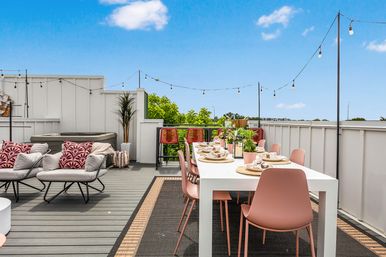 Rooftop patio with a white outdoor dining table and pink chairs set for a meal, lounge seating with patterned pillows, potted plants, hot tub and string lights under a blue sky.