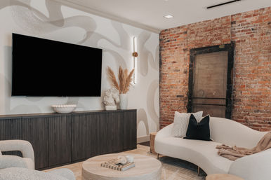 Cozy industrial-chic living room with exposed red brick accent wall, wall-mounted flat-screen TV, dark wood media console, curved white sofa, round wooden coffee table and pampas grass decor.