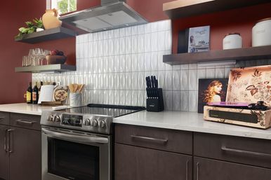 Modern home kitchen with stainless steel stove and hood, white textured oval-tile backsplash, dark wood cabinets, open floating shelves with glassware and cookbooks, knife block and floral record player on white quartz countertops and red accent wall.
