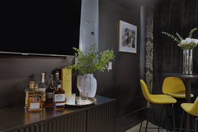 Stylish modern apartment lounge corner with a black console holding assorted liquor bottles, champagne flutes and a vase of greenery on a silver tray, framed wall art and bright yellow bar stools by a small round table.