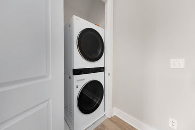 Space-saving modern stacked front-load washer and dryer tucked into a slim laundry closet with light gray walls, white trim and warm wood flooring.