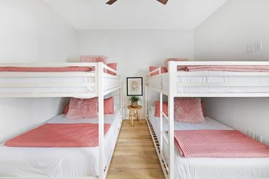 Bright, cozy bedroom with two white twin bunk beds (four beds) dressed in pink quilts and fluffy pillows, light hardwood floors, ceiling fan overhead, and a small stool holding a potted plant and framed art between the beds.